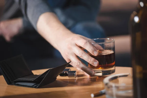 Selektiver Fokus des süchtigen Mannes, der ein Glas Whiskey in der Nähe der leeren Brieftasche auf verschwommenem Vordergrund nimmt — Stockfoto