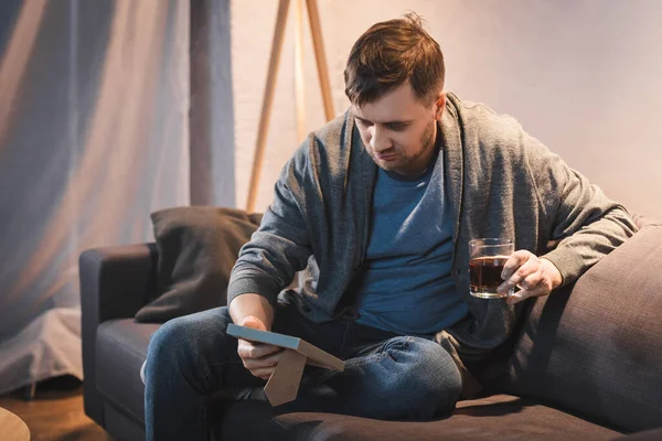 Hombre solitario sosteniendo marco de fotos mientras está sentado en el sofá con un vaso de whisky - foto de stock