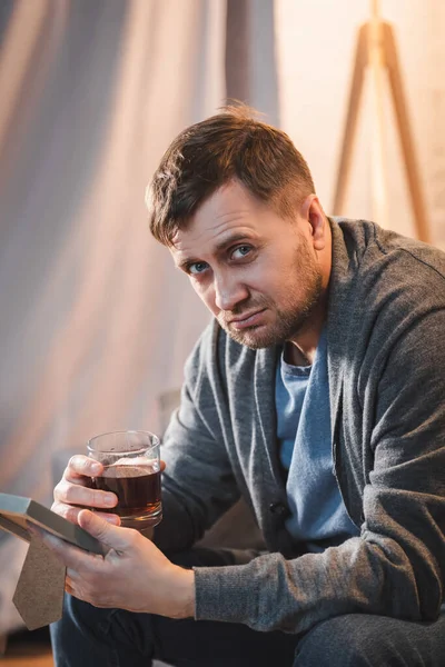 Одинокий человек смотрит в камеру, держа стакан виски и фоторамку — стоковое фото