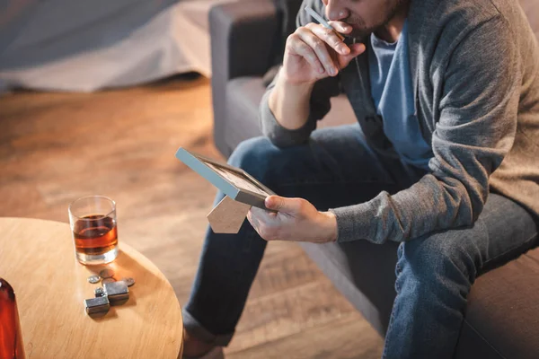 Cropped view of alcohol-addicted man holding photo frame near bottle of whiskey on table — Stock Photo