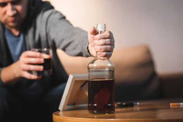 Bêbado homem tomando garrafa de uísque perto de moldura da foto na mesa, fundo borrado — Fotografia de Stock