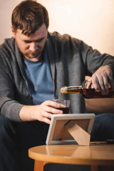 Addicted man pouring whiskey into glass near photo frame, blurred background — Stock Photo