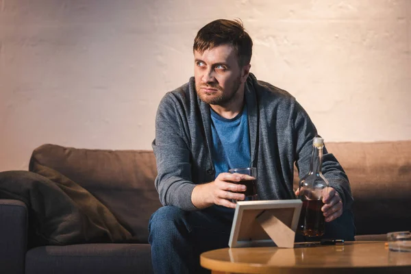 Depressed man holding glass and bottle of whiskey near photo frame on table — Stock Photo