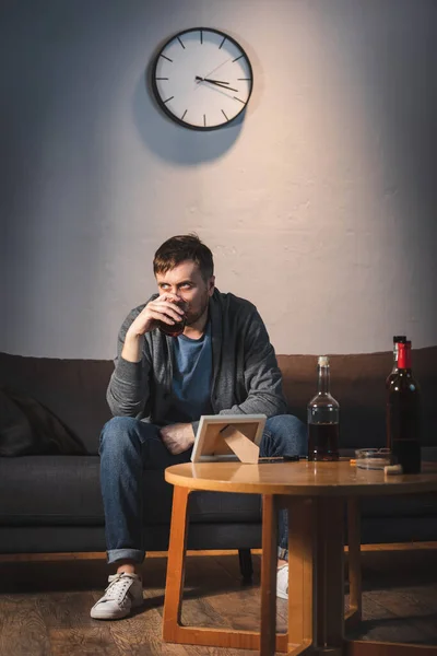 Hombre adicto al alcohol beber whisky cerca de marco de fotos y botellas en la mesa - foto de stock