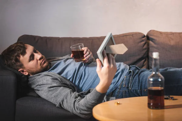 Bouleversé homme couché sur le canapé avec cadre photo et verre de whisky près de la bouteille sur le premier plan flou — Photo de stock