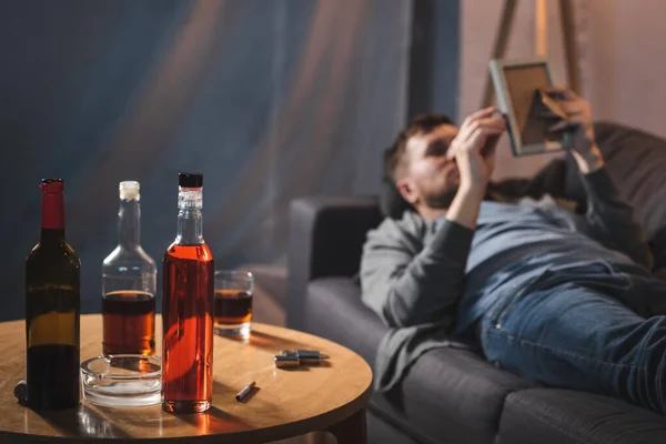 Mesa com garrafas de álcool perto de homem deprimido deitado no sofá com moldura de foto em primeiro plano borrado — Fotografia de Stock