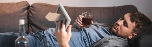 Frustrado hombre acostado en el sofá con un vaso de whisky y marco de fotos cerca de la botella en primer plano borroso, pancarta - foto de stock