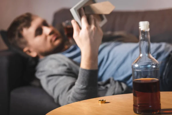 Bouteille de whisky et bague de mariage sur la table près de l'homme déprimé couché sur le canapé avec cadre photo sur fond flou — Photo de stock