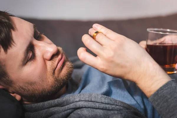 Frustrierter Mann schaut auf Ehering, während er mit Glas Whiskey auf Sofa liegt — Stockfoto