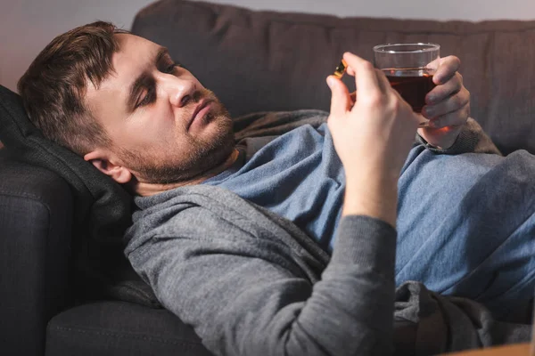 Depressed man holding wedding ring and glass of whiskey while lying on sofa — Stock Photo