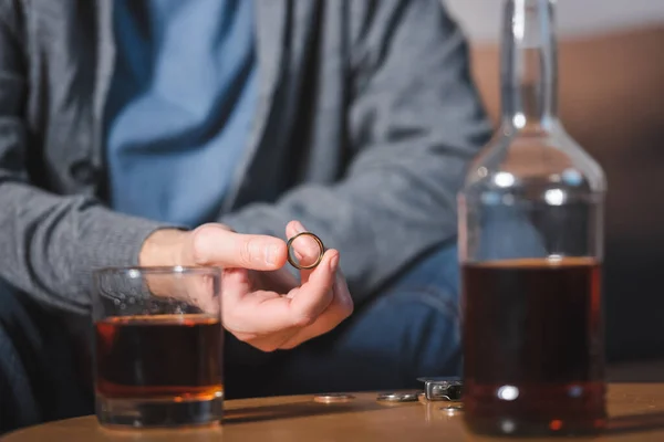 Селективный фокус обручального кольца в руке алкоголика, сидящего рядом со стеклом и бутылкой виски — стоковое фото