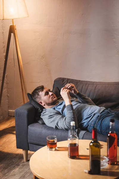 Frustrated man lying on sofa near bottles with alcohol drinks on table at home — Stock Photo