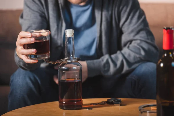 Vista recortada de alcohólico, esposado a la botella, sosteniendo vaso de whisky, fondo borroso - foto de stock