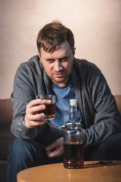 П'яний чоловік, в наручниках до пляшки алкоголю, тримає склянку віскі, розмитий передній план — стокове фото