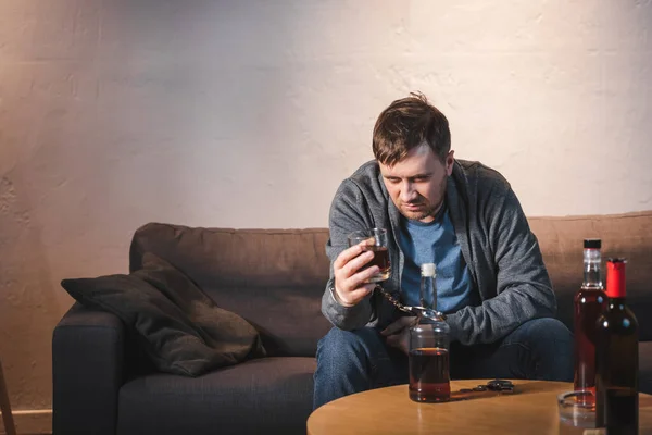 Depressed man, handcuffed to bottle, holding glass of whiskey at home — Stock Photo