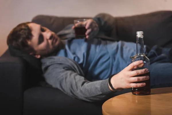 Uomo alcolizzato, ammanettato alla bottiglia, sdraiato sul divano con un bicchiere di whisky — Foto stock