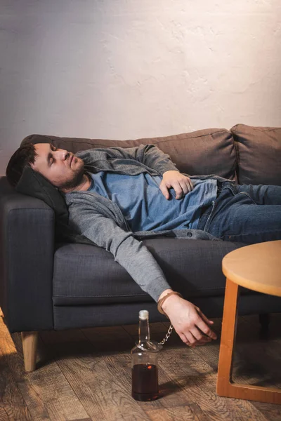 Drunk man, handcuffed to bottle of whiskey, sleeping on sofa at home — Stock Photo