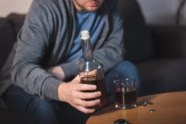 Vista parcial del hombre solitario sosteniendo botella de whisky cerca de vidrio, fondo borroso — Stock Photo
