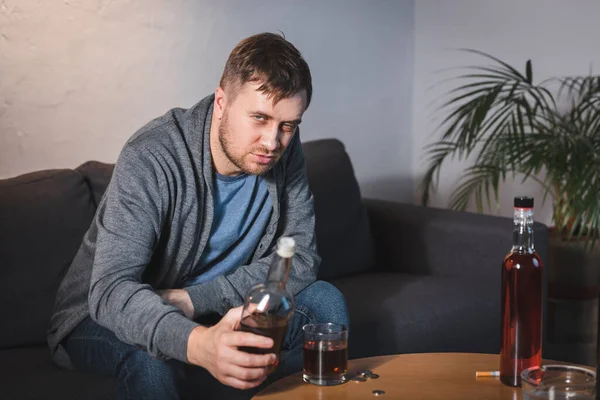 Drunk man looking at camera while holding bottle of whiskey at home — Stock Photo