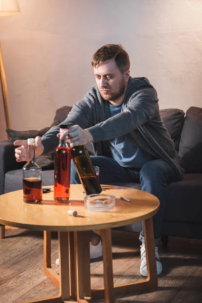 Drunk man taking bottles of alcohol from table while sitting at home alone — Stock Photo
