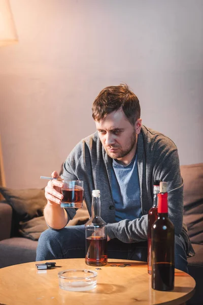 Depresso, uomo solitario che tiene un bicchiere di whisky vicino alle bottiglie sul tavolo a casa — Foto stock