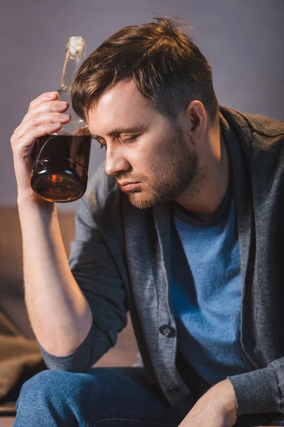 Borracho, hombre deprimido sentado con botella de whisky cerca de la cabeza con los ojos cerrados - foto de stock