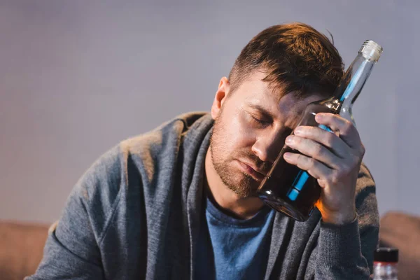 Hombre adicto al alcohol sentado con botella de whisky cerca de la cabeza con los ojos cerrados — Stock Photo