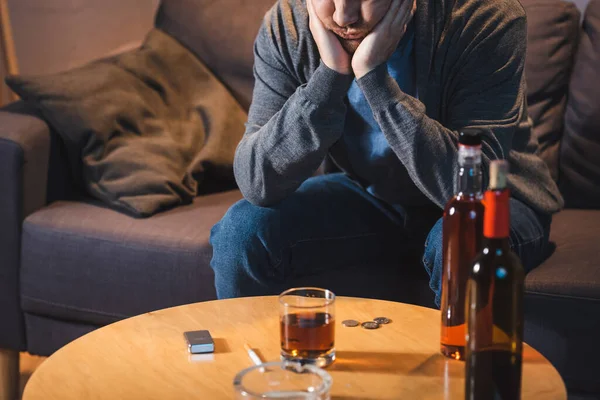 Borracho sentado cerca de la mesa con vidrio y botellas de alcohol en casa - foto de stock