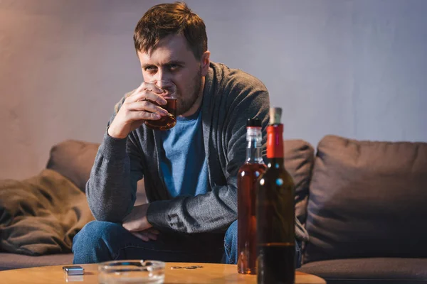 Deprimido, hombre solitario bebiendo whisky cerca de la mesa con botellas de alcohol en primer plano borrosa — Stock Photo