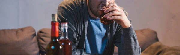 Cropped view of alcohol-addicted man drinking whiskey near bottles on blurred foreground, banner — Stock Photo
