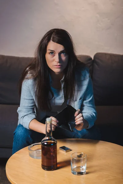 Depressed woman looking at camera while showing empty wallet near bottle of whiskey on table — Stock Photo