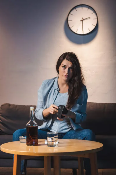 Mujer borracha mirando a la cámara mientras sostiene la cartera vacía cerca de la botella de whisky en la mesa - foto de stock