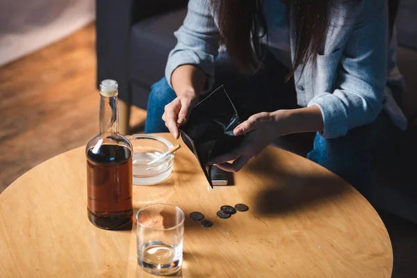 Vista ritagliata della donna che tiene il portafoglio vuoto vicino alla bottiglia di whisky e monete sul tavolo — Foto stock