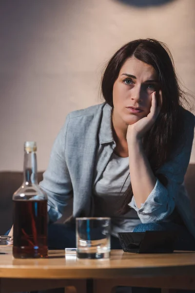 Depressed woman looking away while sitting near bottle of whiskey on blurred foreground — Stock Photo