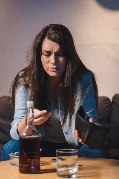 Femme ivre tenant portefeuille vide près du verre et bouteille de whisky au premier plan flou — Photo de stock