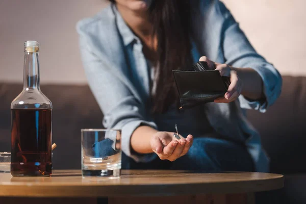 Partial view of woman taking coins from wallet near bottle of whiskey on table, blurred background — Stock Photo