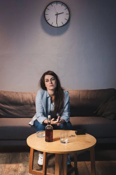 Mulher viciada em álcool frustrado sentado no sofá perto de garrafa de uísque e carteira vazia na mesa — Fotografia de Stock