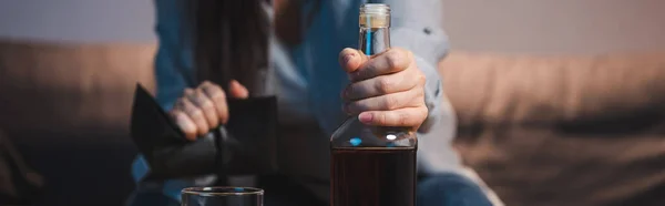 Cropped view of alcoholic woman holding bottle and empty wallet on blurred background, banner — Stock Photo