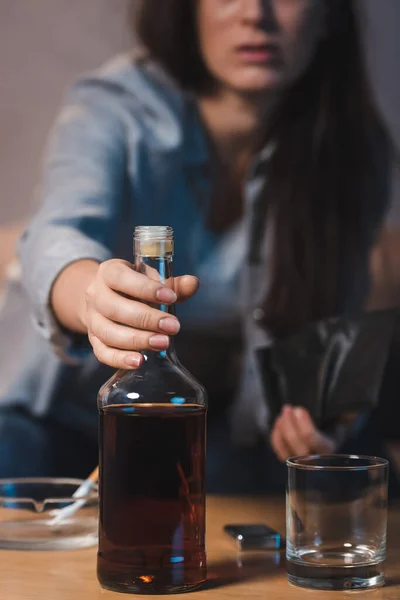 Teilaufnahme einer alkoholisierten Frau, die eine Flasche Whiskey vom Tisch nimmt, verschwommener Hintergrund — Stockfoto