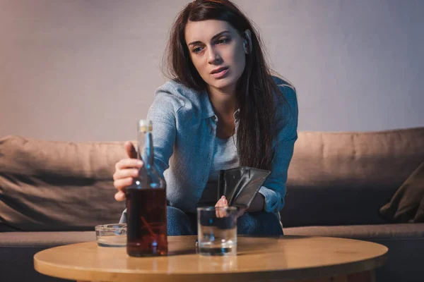 Femme déprimée prenant bouteille de whisky tout en tenant portefeuille vide, avant-plan flou — Photo de stock