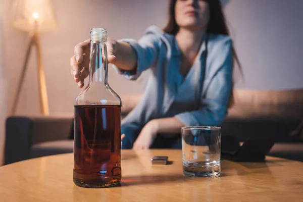 Vista ritagliata di donna alcolizzata che prende bottiglia di whisky da tavolo, sfondo sfocato — Foto stock