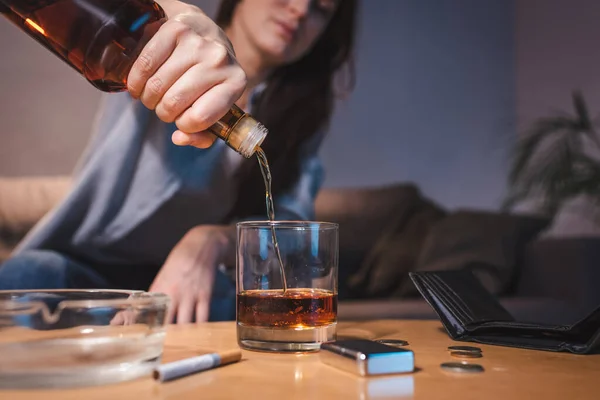 Cropped view of woman pouring whiskey into glass near ashtray, coins and empty wallet, blurred background — Stock Photo