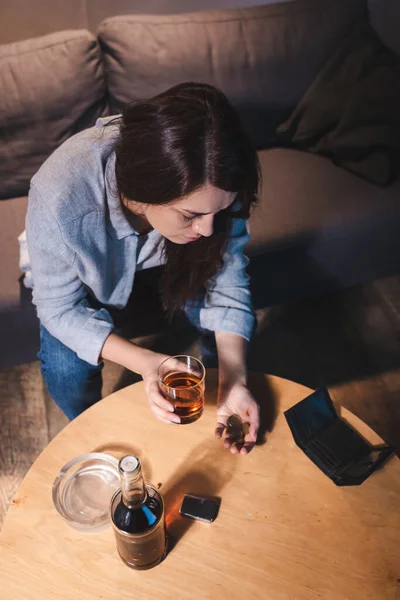 Vista aerea della donna che tiene un bicchiere di whisky e monete vicino alla bottiglia e al portafoglio vuoto — Foto stock