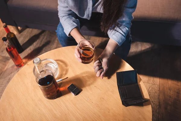 Vue du dessus de la femme alcoolique tenant un verre de cognac et des pièces près du portefeuille vide sur la table — Photo de stock