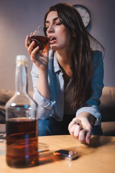 Mujer solitaria bebiendo whisky mientras sostiene monedas cerca de la botella en primer plano borrosa — Stock Photo