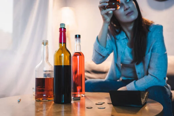 Drunk woman holding glass of alcohol near bottles, coins and empty wallet on table, blurred background — Stock Photo