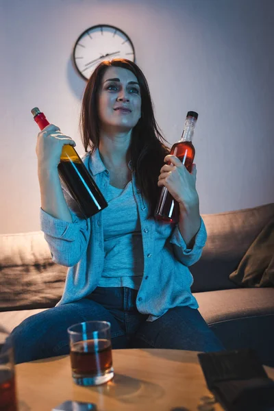 Addicted woman holding bottles of alcohol near glass on blurred foreground — Stock Photo