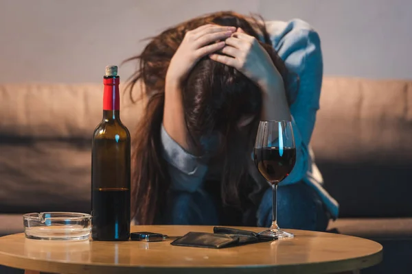 Ivre, femme déprimée assise avec la tête baissée près de la bouteille de vin rouge et portefeuille vide, fond flou — Photo de stock