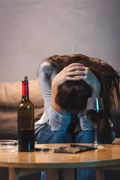 Mujer deprimida sentada con la cabeza inclinada cerca de la botella y el vaso de vino tinto en primer plano borroso - foto de stock