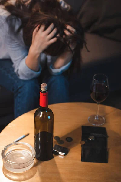 Donna alcolizzata seduta con la testa piegata vicino alla bottiglia di vino rosso, monete e portafoglio vuoto, sfondo sfocato — Foto stock